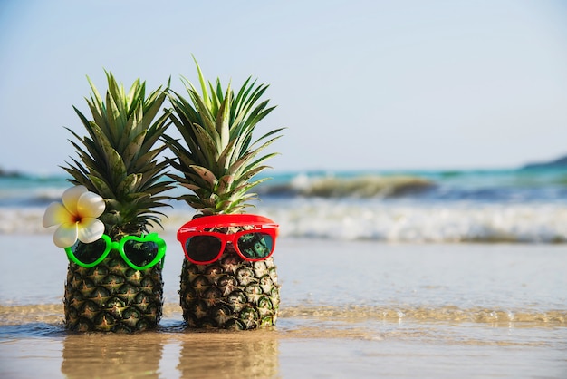 素敵なカップルの新鮮なパイナップルは、海の波のあるきれいな砂のビーチに太陽の素敵なメガネを置きます-海の砂の太陽の休暇のコンセプトを持つ新鮮な果物