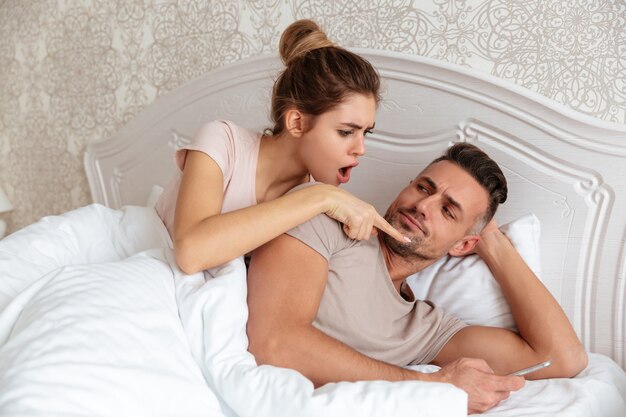 Lovely couple lying together in bed while man using smartphone