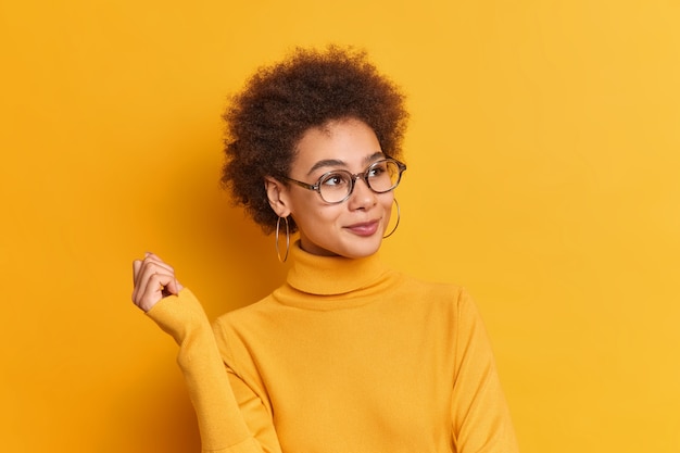 Lovely curly Afro American schoolgirl wears transparent glasses earrings and turtleneck looks dreamy into distance keeps hand raised.