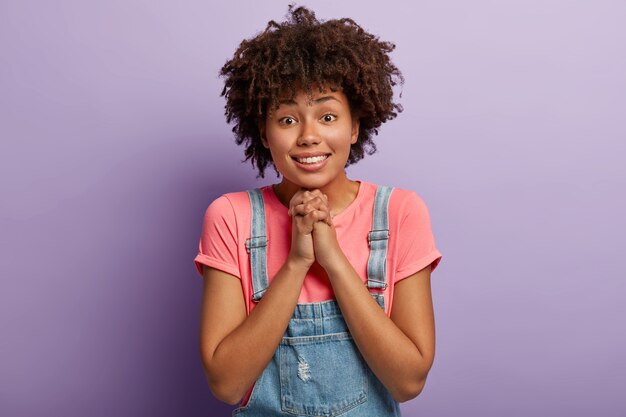 Lovely curly haired girl with angel look, keeps hands pressed together, beggs for favour or help