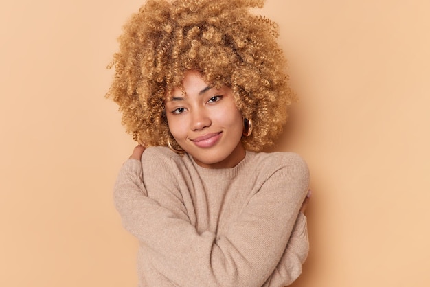 Foto gratuita bella donna dai capelli ricci gode di intimità indossa un nuovo maglione caldo si abbraccia si sente accogliente guarda teneramente alla telecamera sorride dolcemente con amore isolato su sfondo beige. abbracciare il proprio corpo.