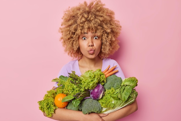 Бесплатное фото Прекрасная кудрявая женщина держит губы сложенными, держит букет продуктов из свежих зеленых овощей, собранных в теплице, выбирает здоровый образ жизни, изолированный на розовом фоне весенние витамины