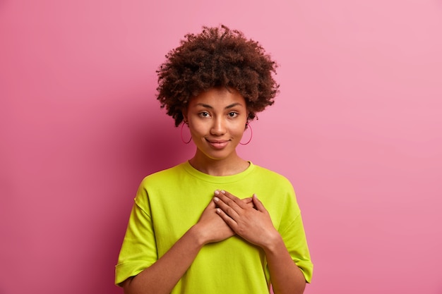 Bella donna dai capelli ricci preme i palmi sul petto, fa un gesto di gratitudine, grazie per l'aiuto o la lode, essendo grata, vestita con una maglietta casual