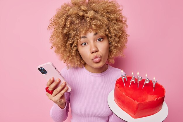 Lovely curly haired young woman keeps lips folded prepares heart cake for husband uses mobile phone for chatting online has romantic expression isolated over pink background Female with dessert