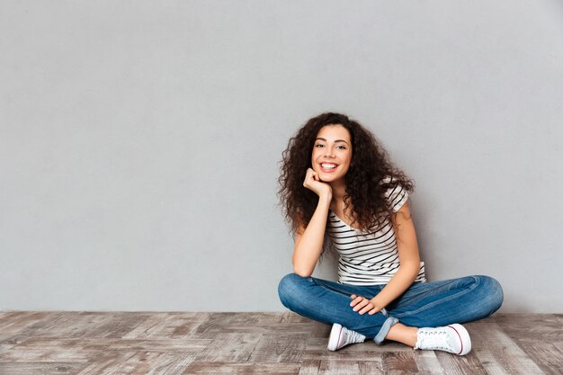 Lovely curly woman in casual clothes sitting in lotus pose on the floor propping up her head with hand being happy and candid over grey wall