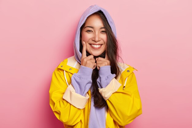 Lovely cute Asain girl wears purple hoody, yellow waterproof raincoat, keeps index finger on cheeks, expresses positive emotions