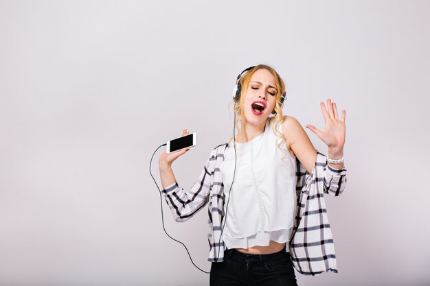 Lovely cute blonde woman enjoying her life, singing and dancing with closed eyes. Mirthful smart girl wearing stylish white blouse with black trousers.