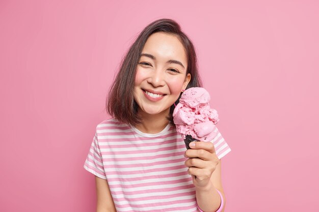Милая темноволосая азиатская женщина любит есть вкусное клубничное мороженое в жаркий летний день, у нее приподнятое настроение, довольное выражение лица, одетая небрежно, позирует с любимым замороженным десертом в помещении