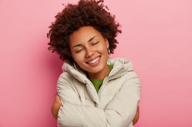 Lovely dark skinned woman embraces herself, enjoys comfort in new winter coat