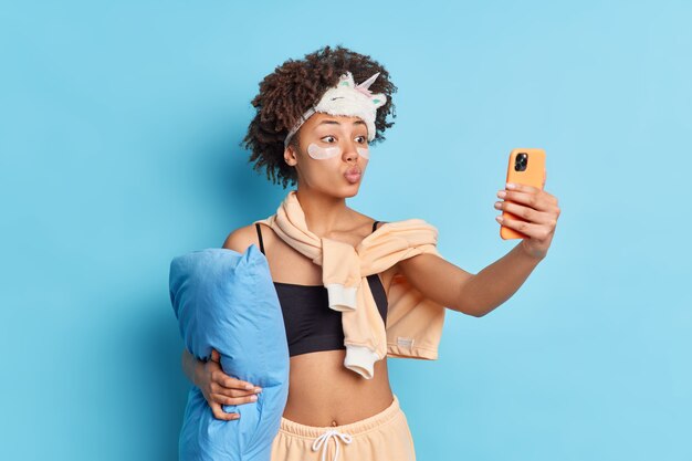 Lovely ethnic woman makes photo of herself keeps lips rounded holds smartphone in front poses in casual domestic pajama sleepmask on forehead isolated over blue background. Selfie before sleep