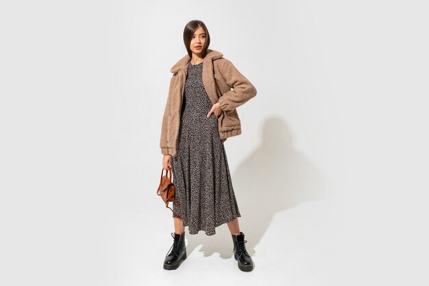Lovely european model in stylish fur coat and dress . Wearing ankle boot in black leather. Holding brown handbag.