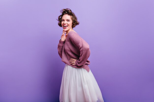 長い白いスカートでポーズをとるスタイリッシュなヘアカットで素敵な興奮した女性。明るい紫色の壁に分離された楽しいブルネットの女性モデルの屋内写真。
