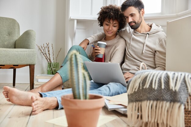 Lovely family couple cuddle together, dressed casually, enjoy domestic atmosphere, synchronize data on laptop computer, work on family business project, drink hot beverage, cactus in foreground