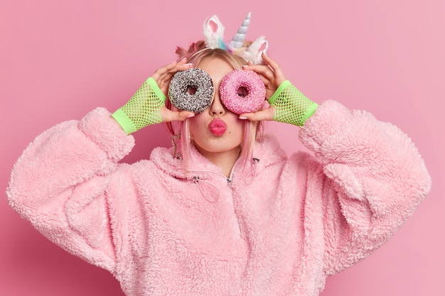 Free photo lovely fashionable teenage girl holds glazed sweet doughnuts over eyes keeps lips rounded