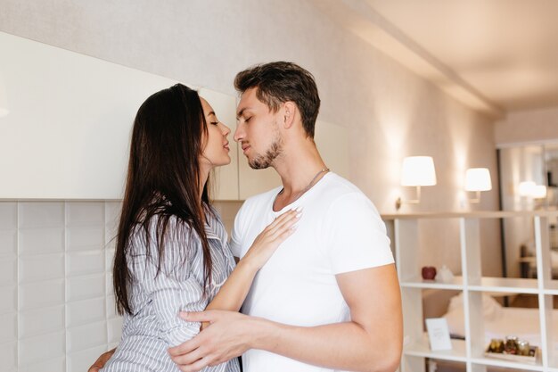 Lovely female model with long dark hairstyle embracing with caucasian man in modern kitchen