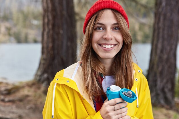 Lovely female tourist has hot drink from thermos in spring forest, wears red hat and yellow raincoat, smiles broadly at camera