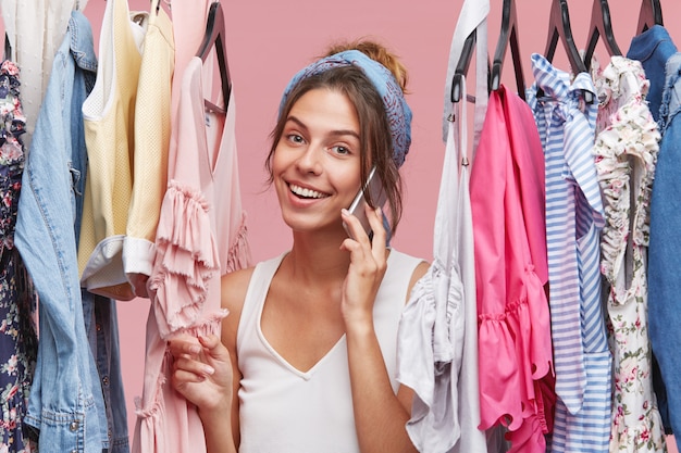 Lovely female wearing scarf on head and casual T-shirt, having good mood while speaking with someone over cell phone, standing near rack of clothes