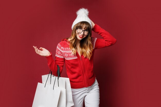 Милая девушка в белой шерстяной шапке и красном зимнем свитере позирует с сумками