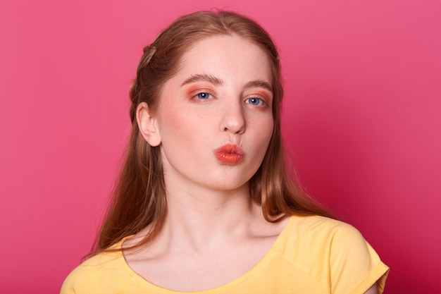 lovely female with long straight hair, wears yellow t shirt, rounds lips as going to recieve kiss from boyfriend, has mysterious facial expression, isolated on pink.