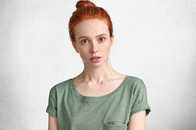 Lovely freckled woman with ginger hair knot, dressed casually, listens attentively lecture, isolated over white