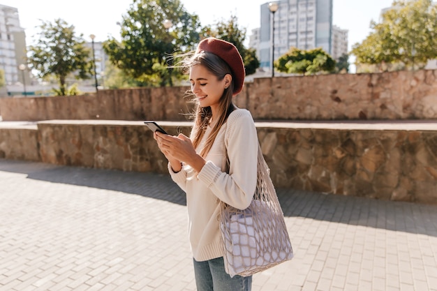 Lovely french girl with bag standing on the street in sunny autumn day. Jocund lady in casual attire texting message.