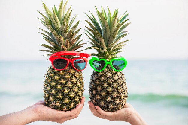 Lovely fresh couple pineapples putting glasses in tourist hands with sea wave - happy love and fun with healthy vacation concept