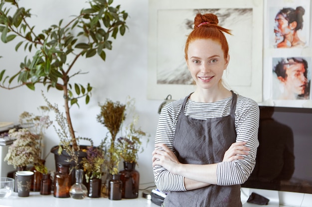 Lovely ginger female studying art in workshop, smiling gently, standing crossed hands against beautiful pictures.