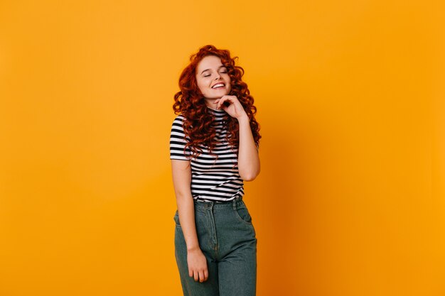 Lovely ginger girl with wavy hair touches her chin and laughs with her eyes closed, posing in jeans on orange space.