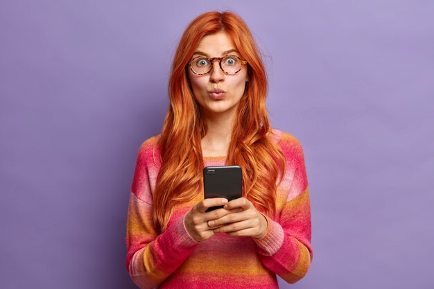 милая рыжая женщина выглядит удивительно, держит губы сложенными, использует современный смартфон
