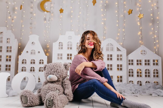 Милая девочка 25 лет сидит на полу со своей плюшевой игрушкой над белой стеной с теплыми украшениями, такими как яркие золотые звезды