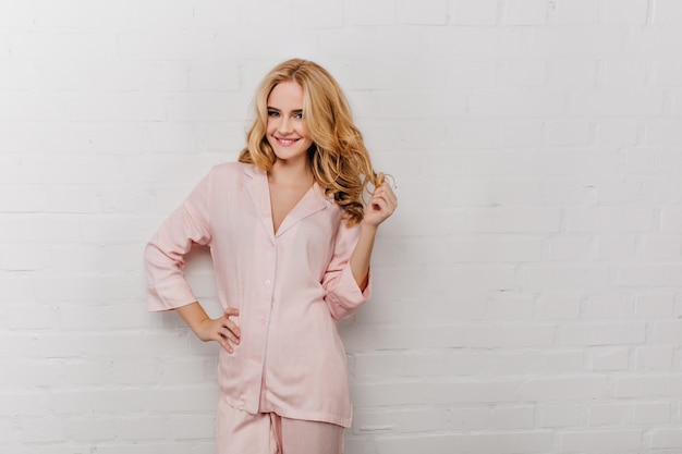Lovely girl in cotton pyjama playing with her wavy hair.  portrait of laughing woman in pink night-suit smiling on white wall.