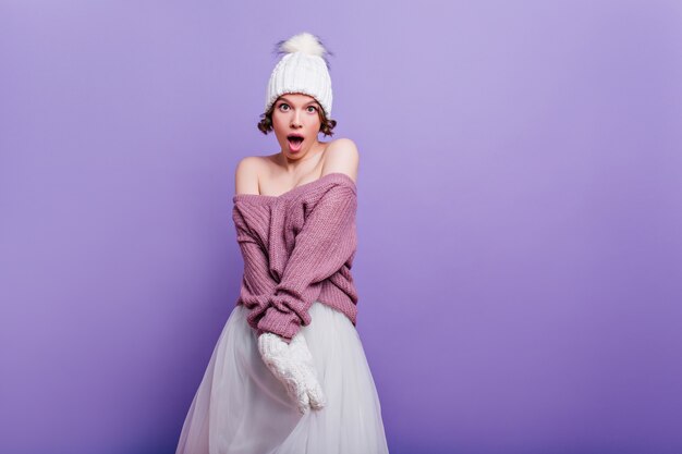 Lovely girl in purple sweater expressing amazement isolated on bright wall. Surprised short-haired brunette woman in white skirt standing 