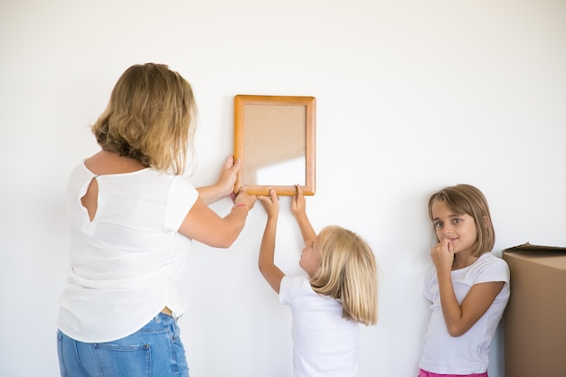 Lovely girl putting frame on white wall with help of mom