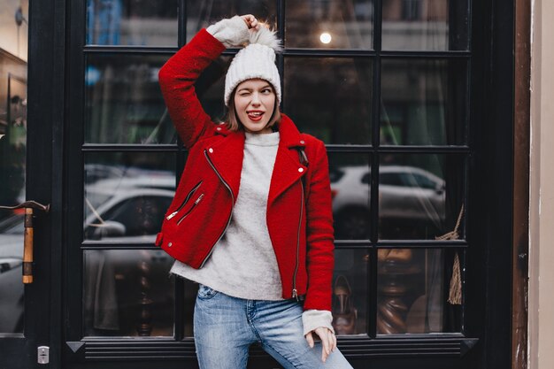 Милая девушка в красном пальто и джинсах веселится, подмигивая, показывая язык и держа помпон вязаной шапки напротив окна.
