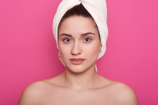 Free photo lovely girl wearing white towel on head, posing with bared shoulders isolated on pink studio, attractive lady looking directly at camera, having clean perfect skin. skinncare concept.