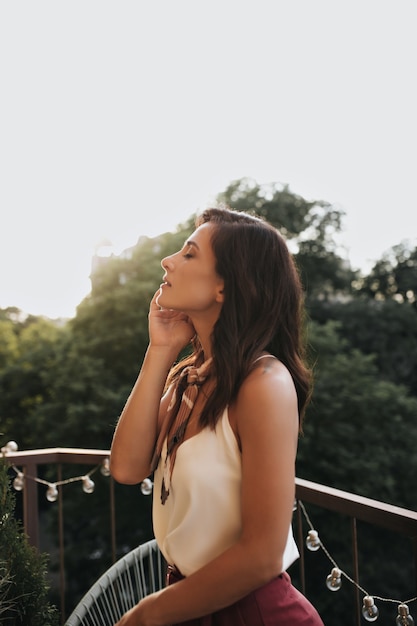 Lovely girl in white top and silk scarf enjoys summer day on balcony. profile photo of beautiful woman in stylish outfit on terrace.