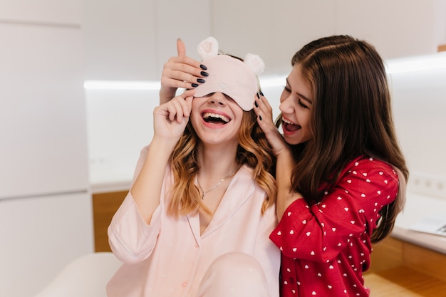 Lovely girl with black manicure playing with her sister in morning. Funny curly female model in pink sleepmask relaxing with best friend.