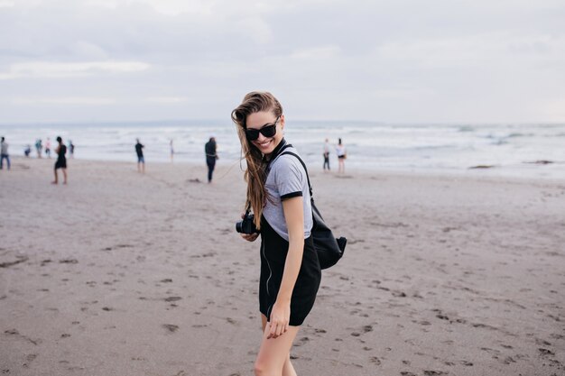 Милая девушка с камерой, проводящей время на песчаном пляже в пасмурный день. Наружная фотография приятной женщины-фотографа, смеющейся в выходные на море.