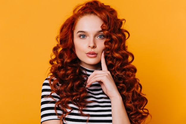 Lovely girl with red curls and blue eyes touches her cheek with her finger and looks thoughtfully at camera on isolated orange space.