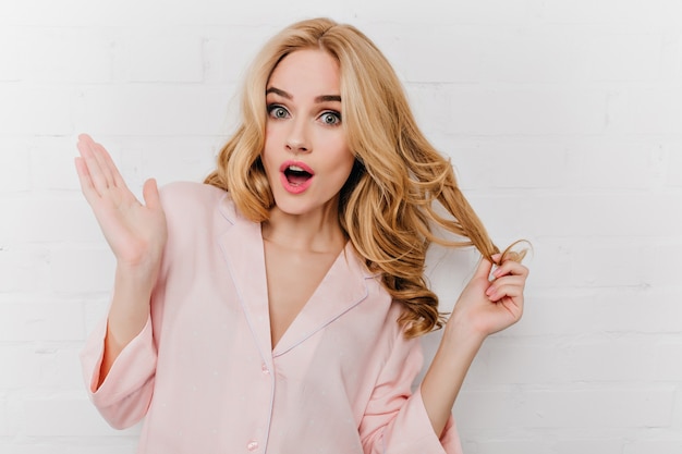 Lovely girl with shiny wavy hair expressing amazement on white wall. Photo of emotional european woman in pink cotton pajamas.