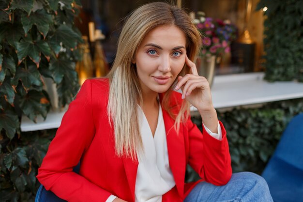 Lovely good- looking blond woman in red jacket posing in city cafe