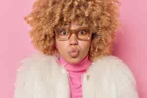 Free photo lovely good looking woman with curly bushy hair has romantic expression confesses in love keeps lips rounded wears spectacles white fur coat isolated over pink background blows mwah at camera