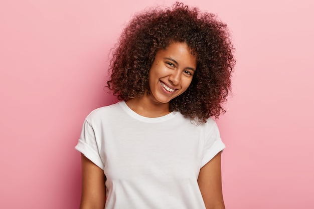 Bella donna bellissima con pelle sana, capelli ricci e folti, inclina la testa, sorride felice, ha poco spazio tra i denti, esprime buone emozioni, ama posare, indossa una maglietta bianca casual