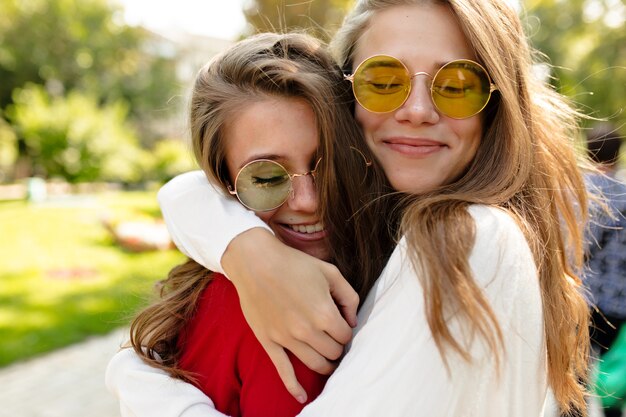 Lovely happy girls walking outside in sunny day. beautiful lovely woman in bright glasses hugging her friend and closed eyes with great smile, best friends, sisters, posivite mood