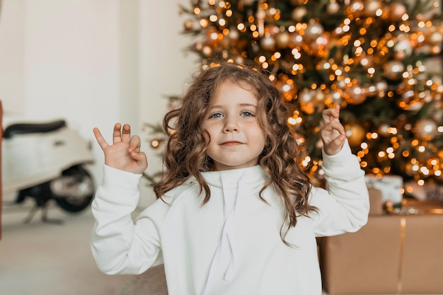カールの服を着た白いニットの服を着て、クリスマスツリーの前で手を上げて笑っている素敵な幸せな少女