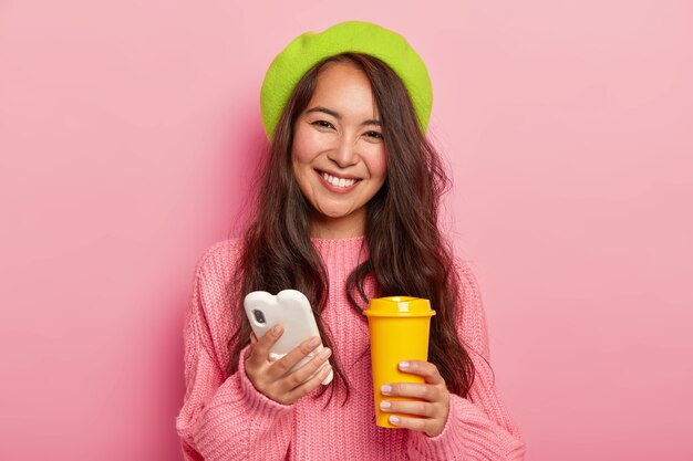 기쁜 표정으로 사랑스러운 행복한 여자, 소셜 네트워크를 서핑하고 온라인 채팅을 위해 핸드폰을 사용하고 커피와 함께 노란색 테이크 아웃 컵을 보유하고 있습니다.