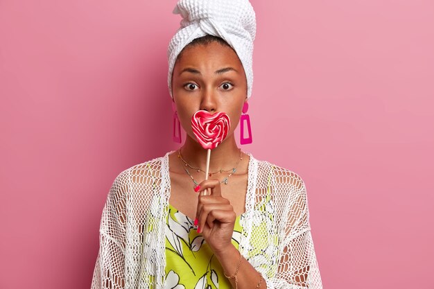 Lovely healthy woman covers mouth with bright heart shaped lollipop, going to satisfy her sweet tooth, wears domestic clothes and jewelry, isolated on pink wall. Female with delicious candy