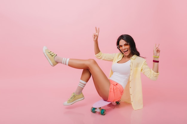 Lovely hispanic woman with bronze skin in denim shorts having fun during indoor summer. Good-looking asian girl with longboard spending free time.