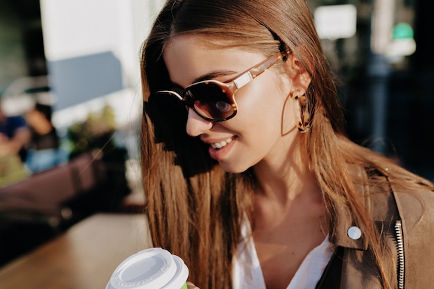 Lovely lady with dark hair wearing sunglasses drinks coffee on wooden outdoor terrace with golden leaves on background. Outdoor portrait of gorgeous white female model in the city