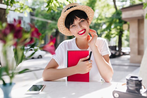 Lovely lady with happy face expression holding red notebook resting in outdoor cafe with phone